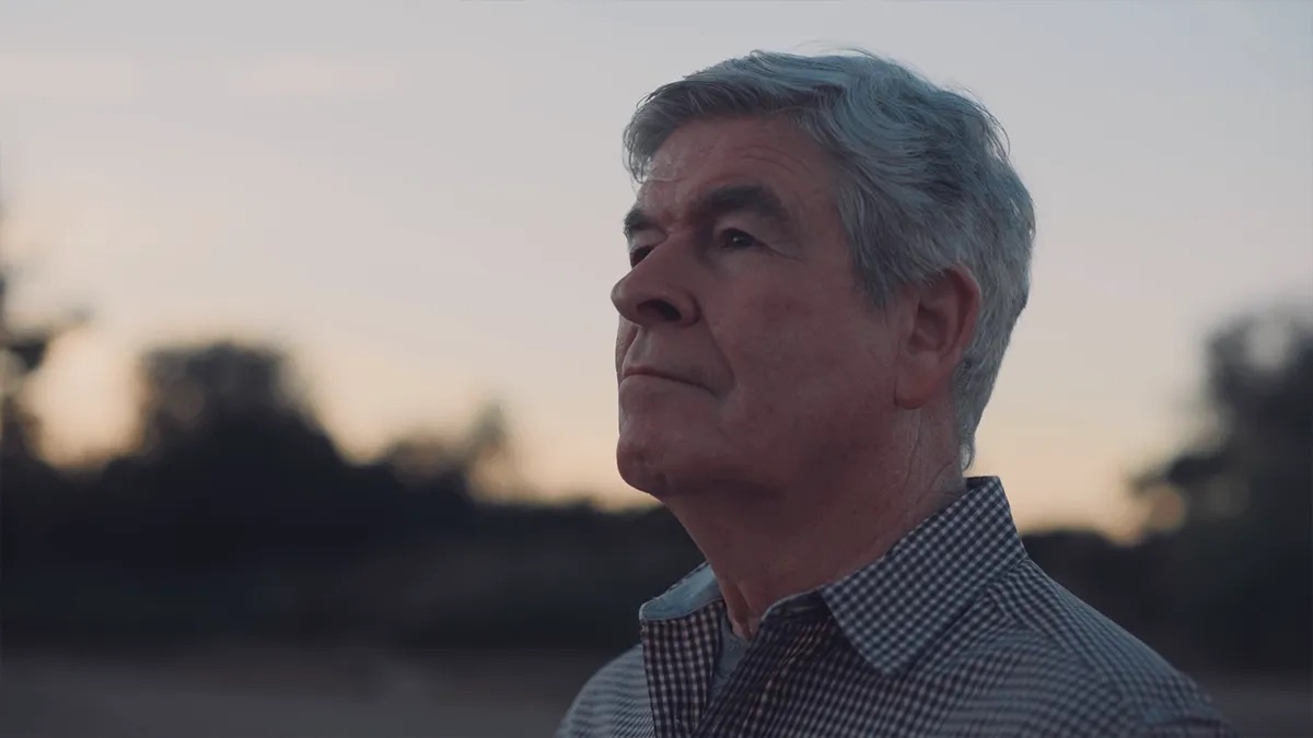 John Taylor attorney profile thumbnail. Profile shot at dusk, a man in a check button-down shirt with tree branches behind him.