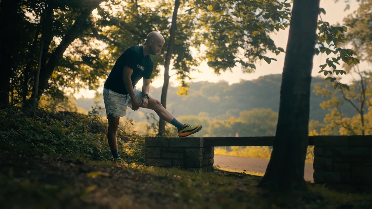 Jonathan Bobbitt attorney profile thumbnail. Stretching his right leg on a stone bench outdoors, a man in a black T-shirt and patterned shorts with wooded hills behind him.