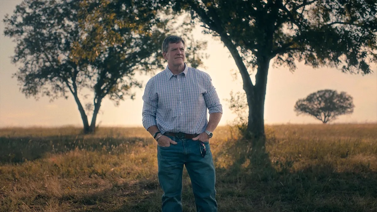 Marcus Mears attorney profile thumbnail. Dry grass field with trees in the background, a man in a plaid button-down shirt and jeans.