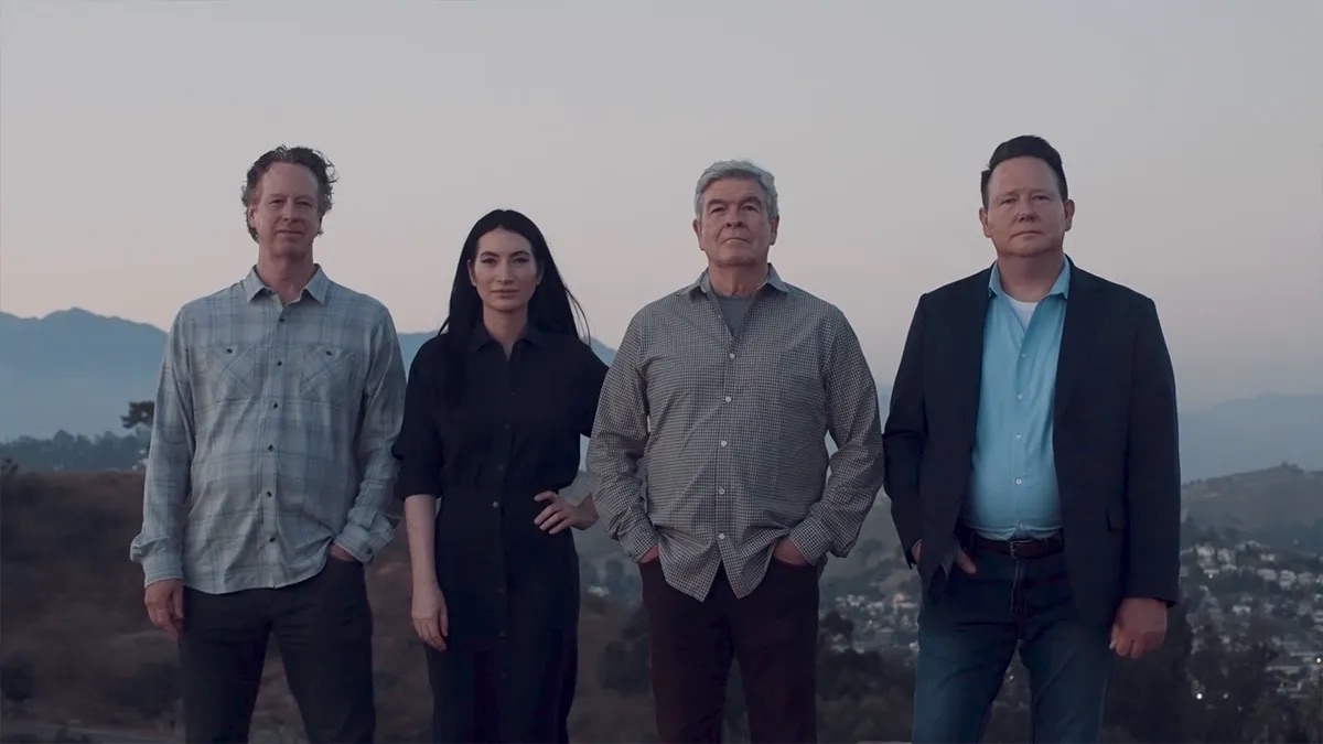 Four people standing side by side on a hillside at dusk, with the houses and hills of Los Angeles in the distance