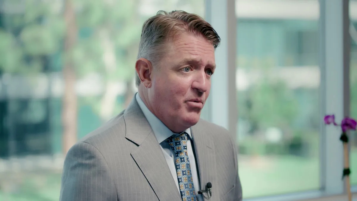 KMD Law practice area video thumbnail on herpes transmission lawsuits. Close shot of an attorney in a gray pinstripe suit beside office windows, a small mic clipped to his tie.