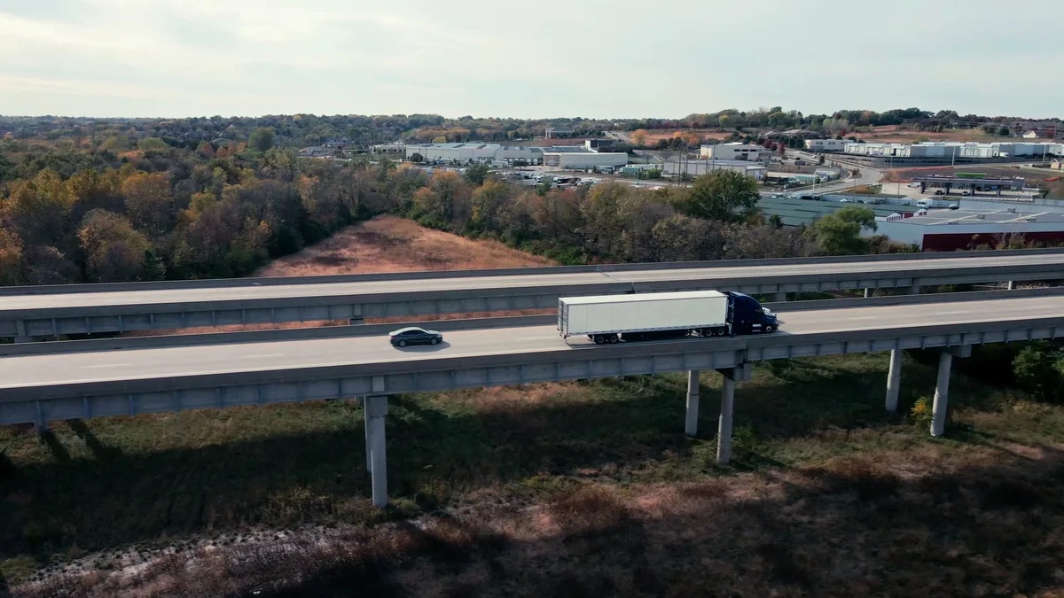 Kevin Patrick Law practice area video thumbnail on truck accidents. Elevated view of a highway with a semi truck and a car crossing side by side, trees and industrial buildings in the background.
