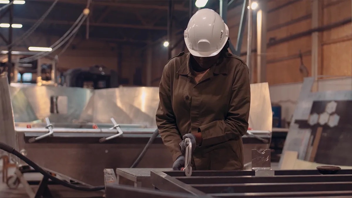 Practice area video thumbnail on products liability. A person in a white hard hat and brown coveralls works with metal in an industrial shop, overhead lights and equipment behind them.