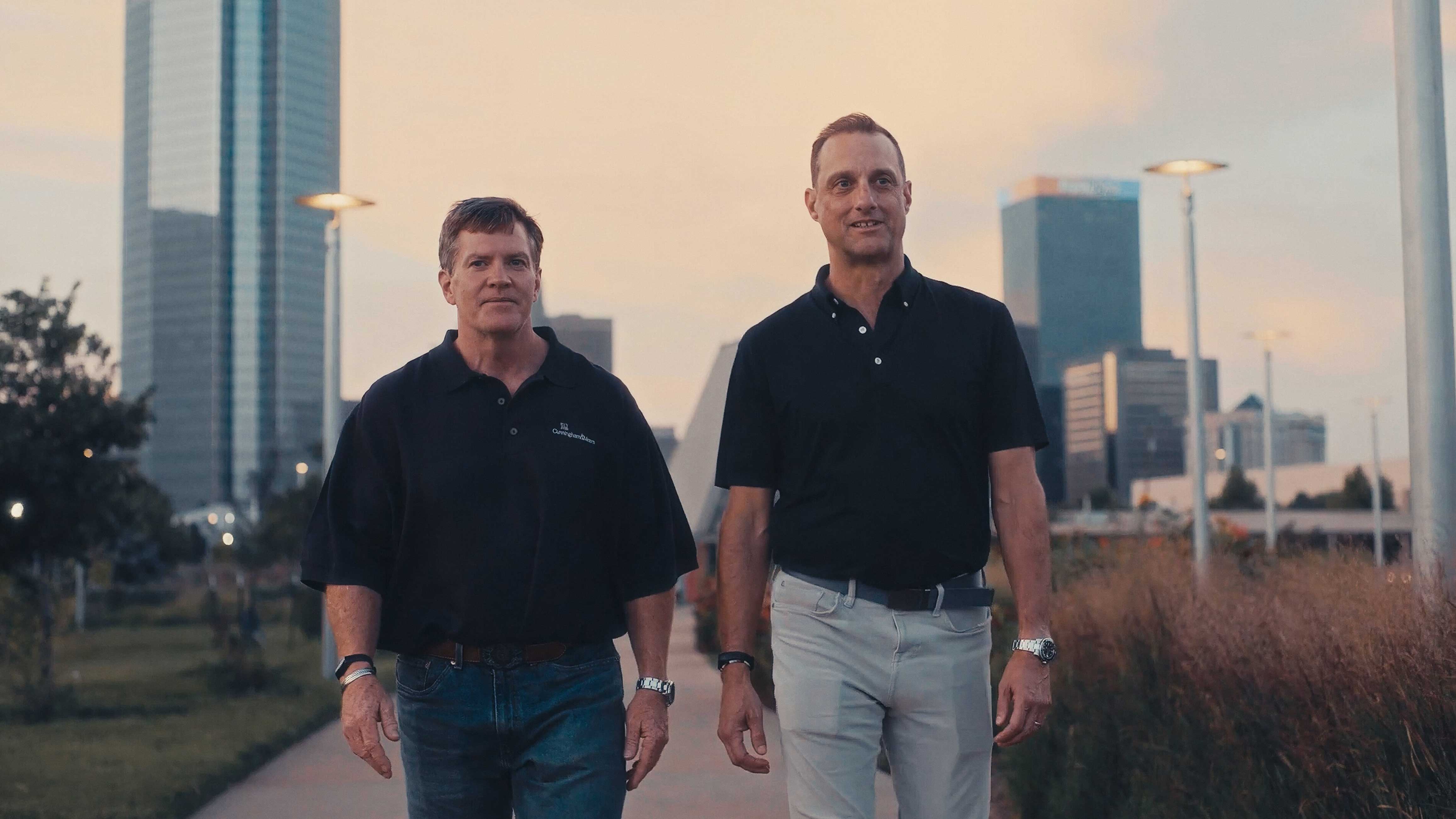 Two men in dark polo shirts walking toward the viewer at nightfall, street lamps lit and the downtown Oklahoma City buildings behind them