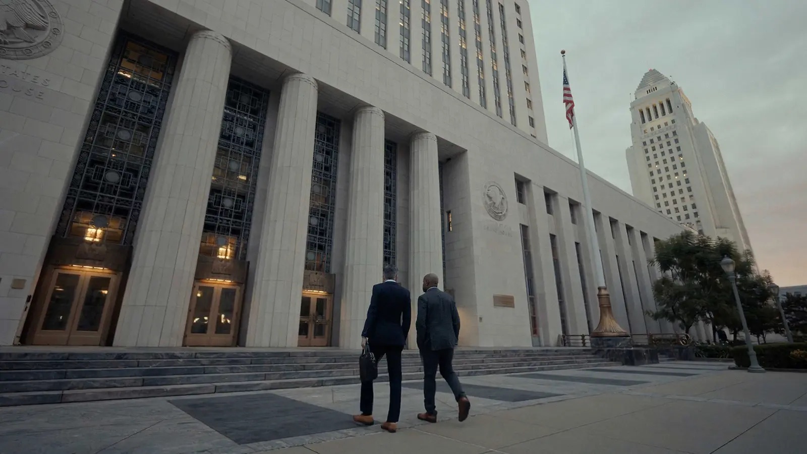 Two suited men heading toward the United States Courthouse in downtown Los Angeles at dusk, LA City Hall rising behind them