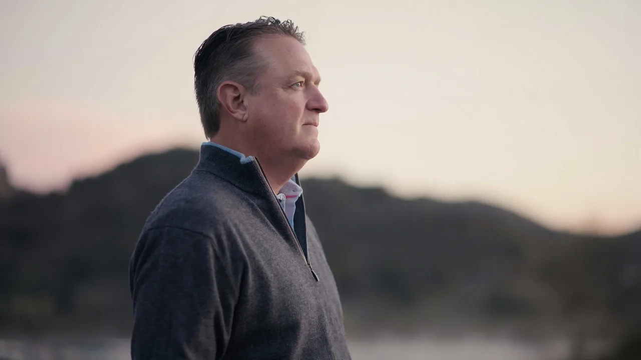 Keith Davidson of KMD Law, grey-haired in a grey quarter-zip, photographed in profile at dusk with soft hills and water behind him