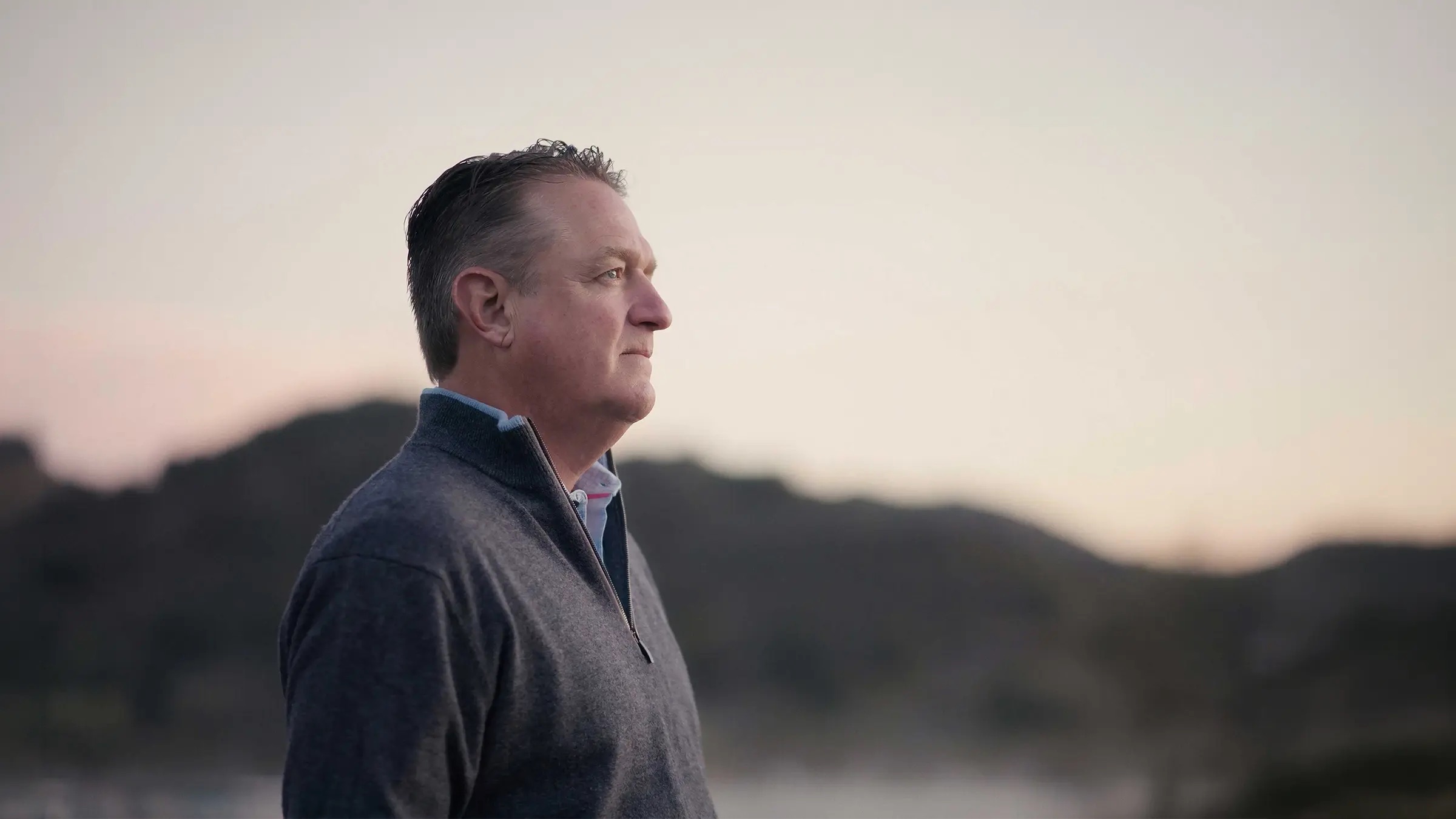 A man in profile wearing a grey quarter zip, hills and water blurred behind him