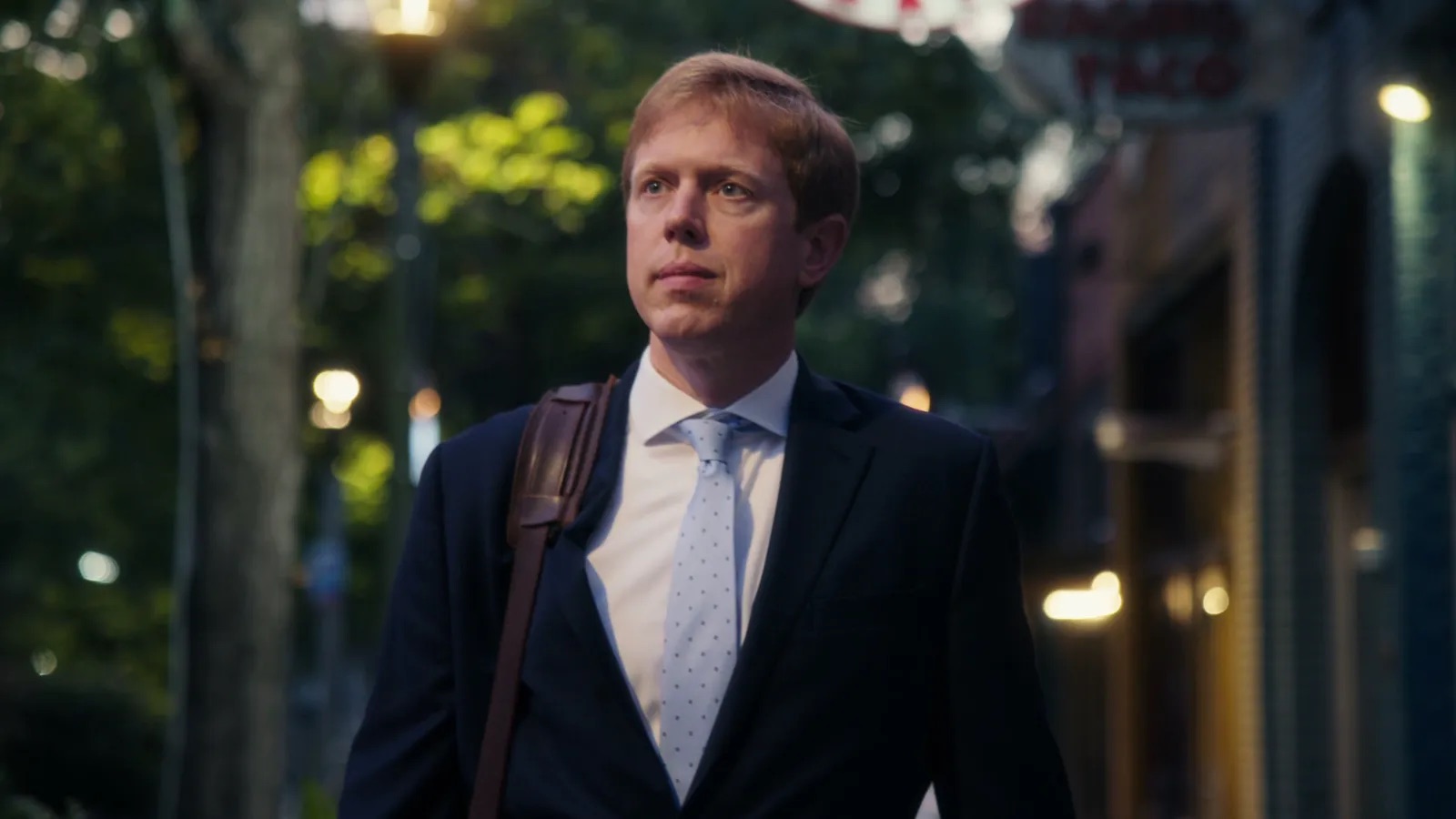 A man in a navy suit walking a city street at dusk, bag strap over his shoulder