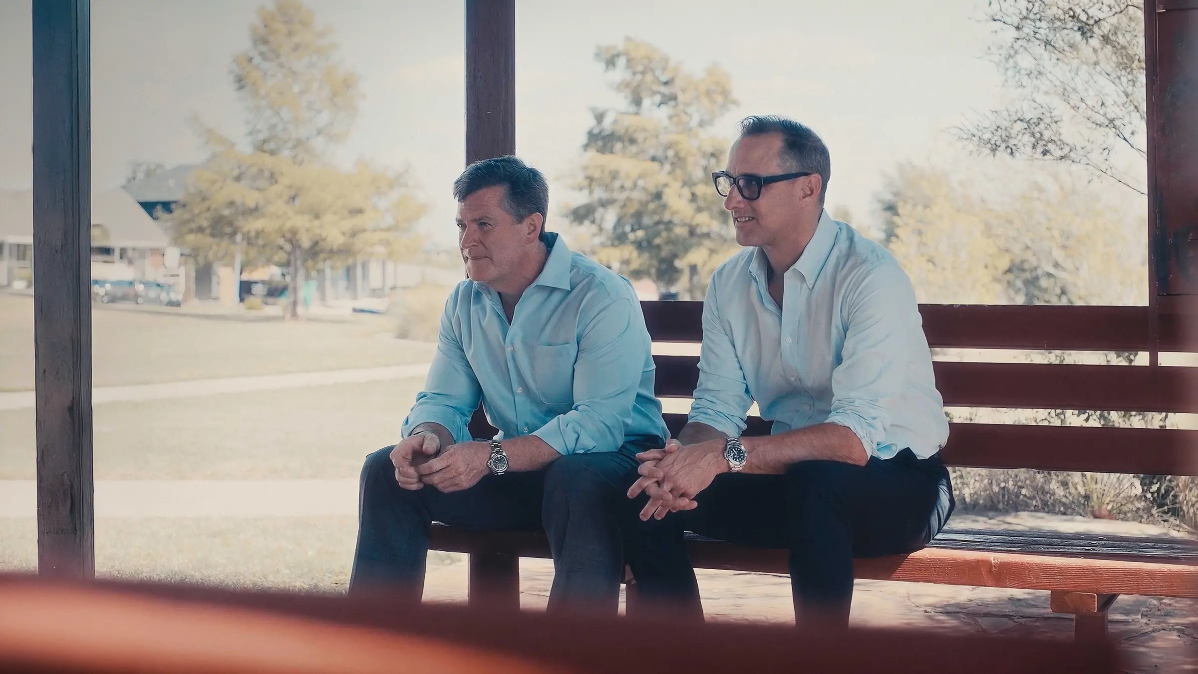 Two men in light blue shirts sitting on a bench outdoors, looking off camera