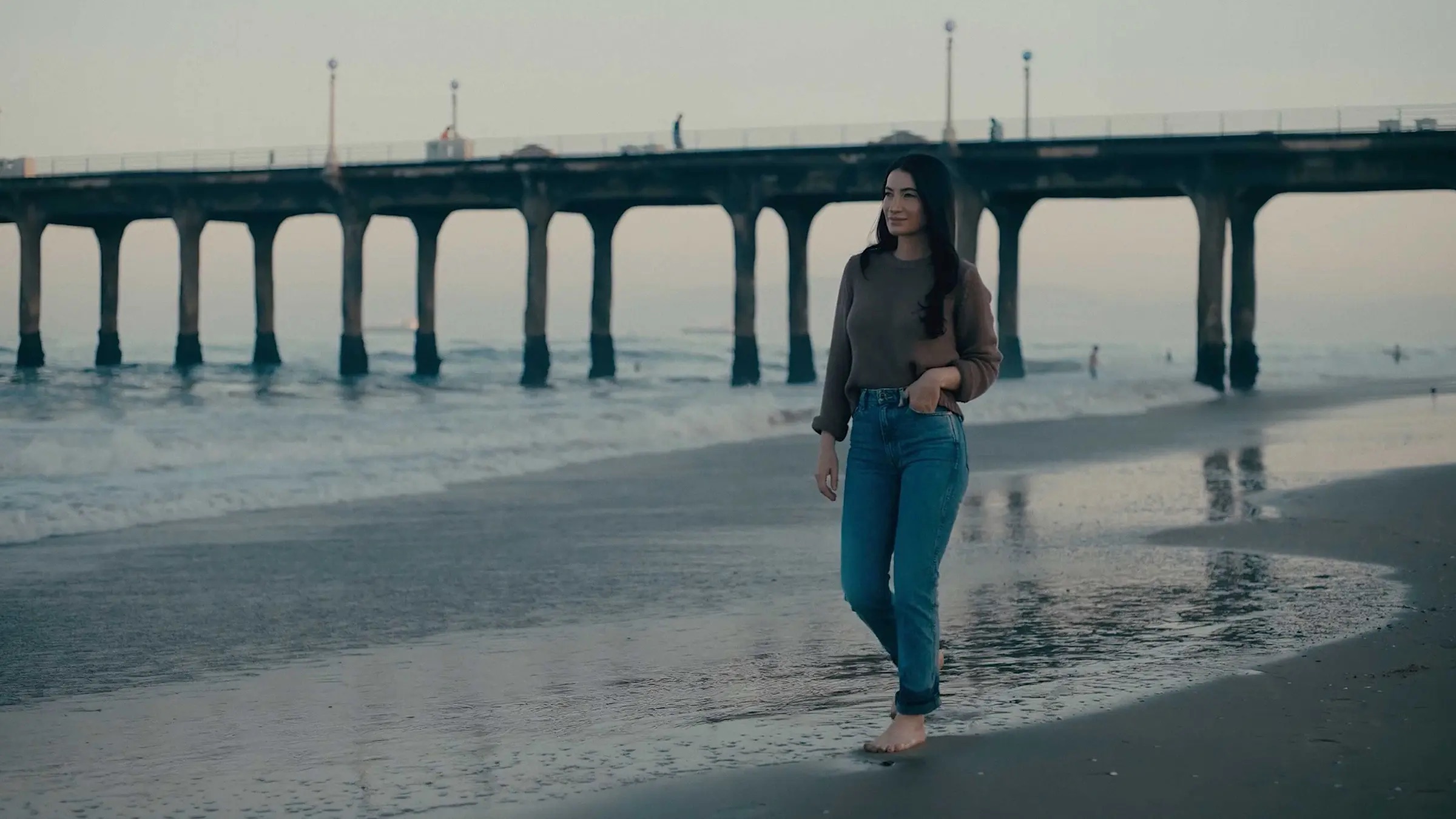 A woman walking barefoot on wet sand beside a pier at dusk